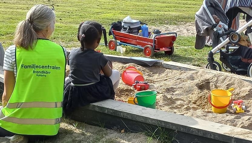 Utomhus i lekpark. Barn leker i sandlåda. Pedagog i varselväst. Skrinda med matsäck.