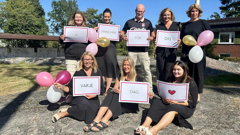 Gruppbild på personal som jobbar med trygghet. De håller i var sitt papper som bildar ordet, som bilder Vi finns här för dig varje dag