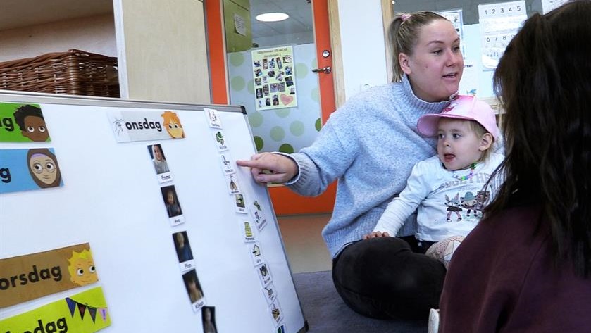 Förskolepedagog sitter med ett barn i knä och pekar på en tavla med små bilder på Förskolepedagog sitter med ett barn i knä och pekar på en tavla med små bilder på