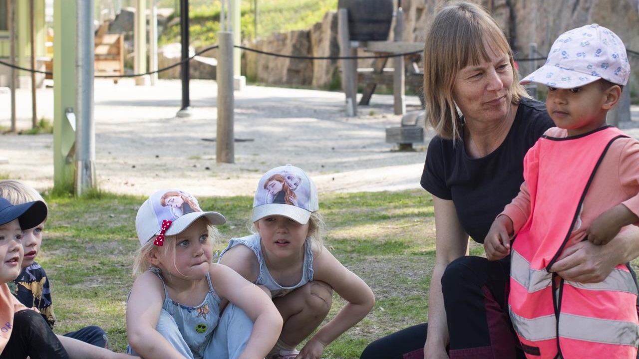 En förskolepedagog står på knä vid ett barn och håller om och tittar på det. Några barn sitter bredvid En förskolepedagog står på knä vid ett barn och håller om och tittar på det. Några barn sitter bredvid