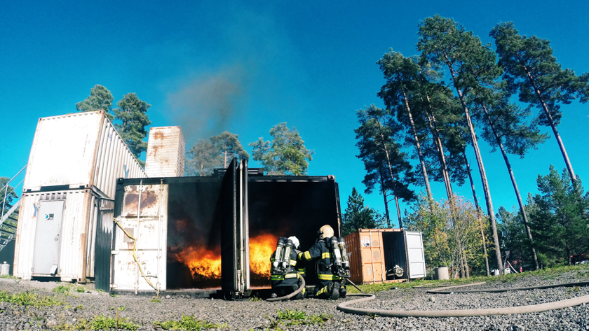 En brinnande övningscontainer och två brandmän utanför En brinnande övningscontainer och två brandmän utanför
